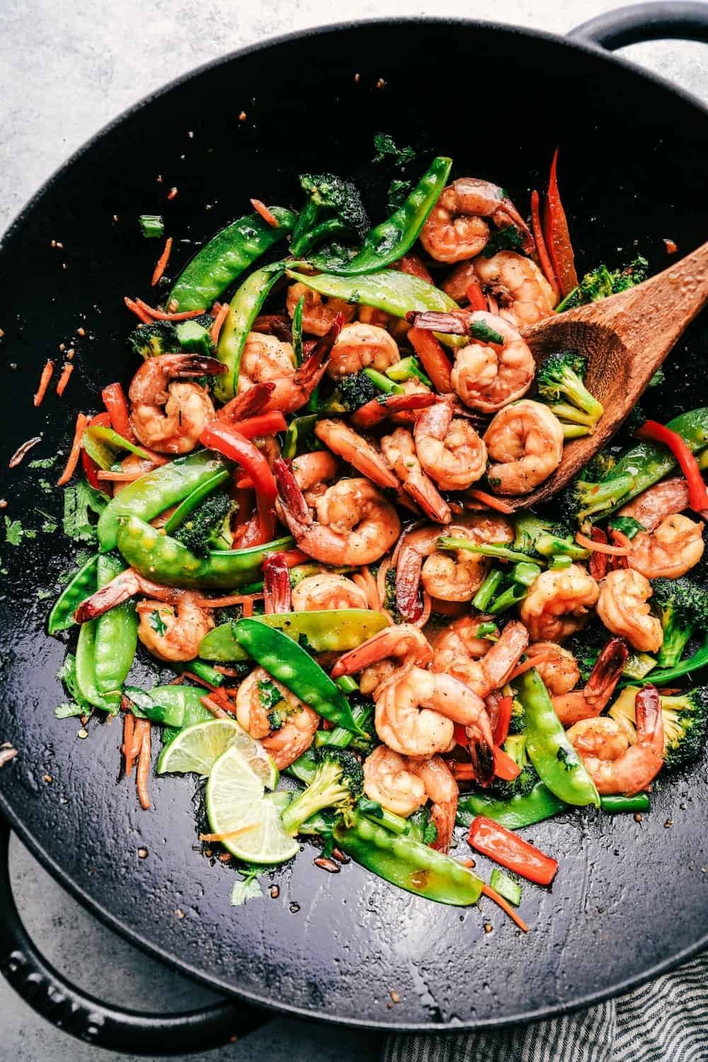 Shrimp stare in a large wok with lime slices and a wooden spoon.